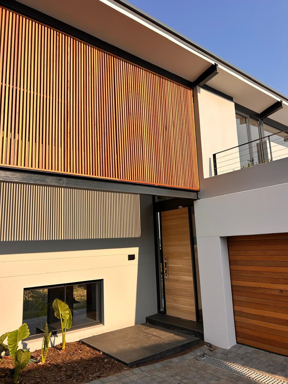 Timber pivot front door beneath a vertical timber slat screen, beside a cedar garage door
