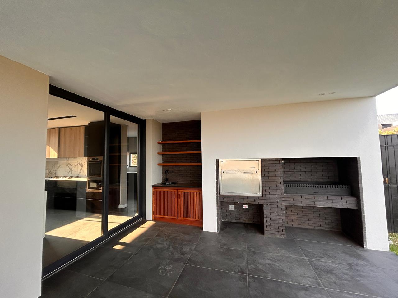 Covered patio with a built-in brick braai, timber shelving and a sink, opening onto the kitchen